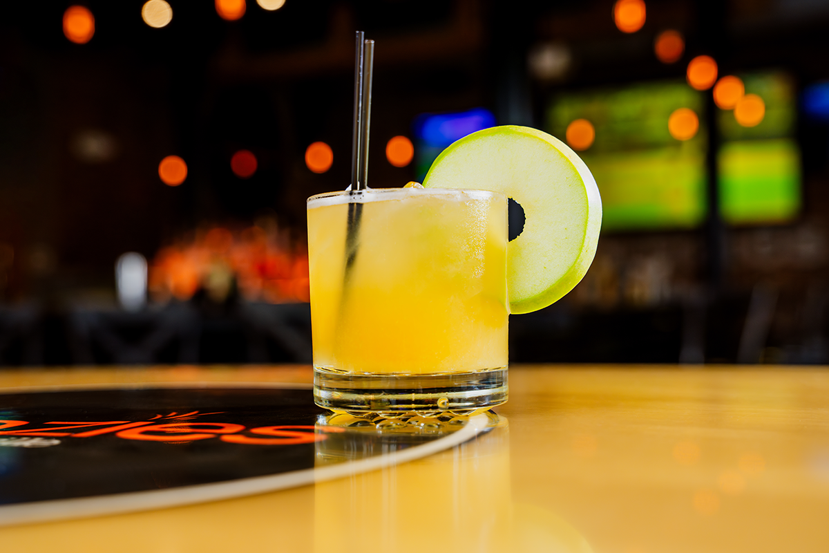Cocktail in a glass with apple slice garnish and straw on a bar, orange-toned lighting.