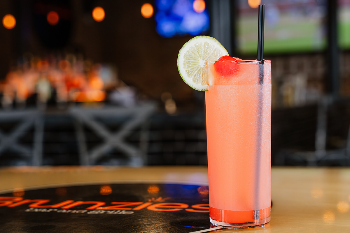 Pink cocktail in tall glass with lime slice and cherry on bar.