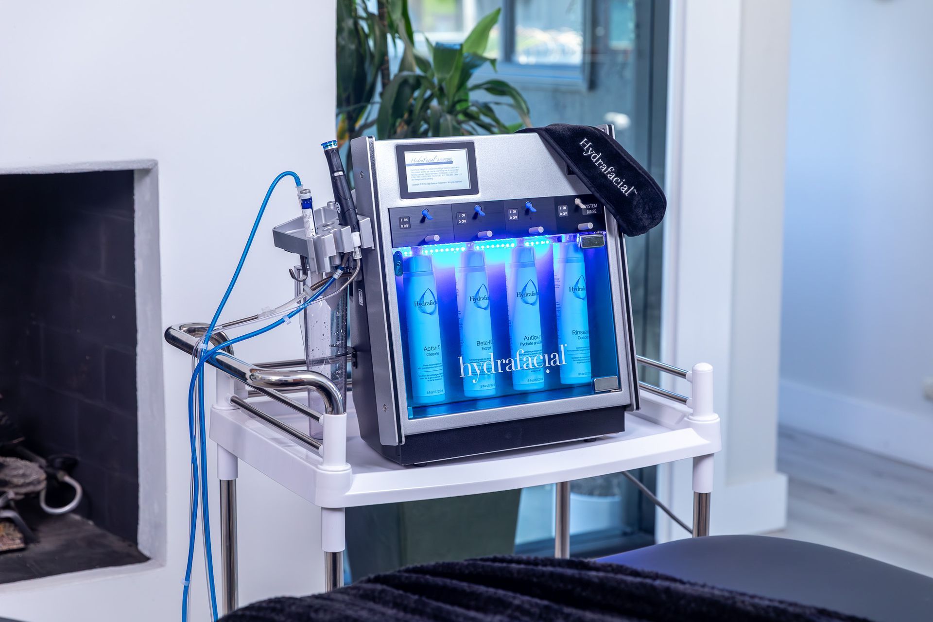 HydraFacial machine on a rolling white cart in a bright room; blue liquid in the chambers.