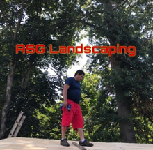Man in red shorts near RSG Landscaping logo, outdoors, looking down.