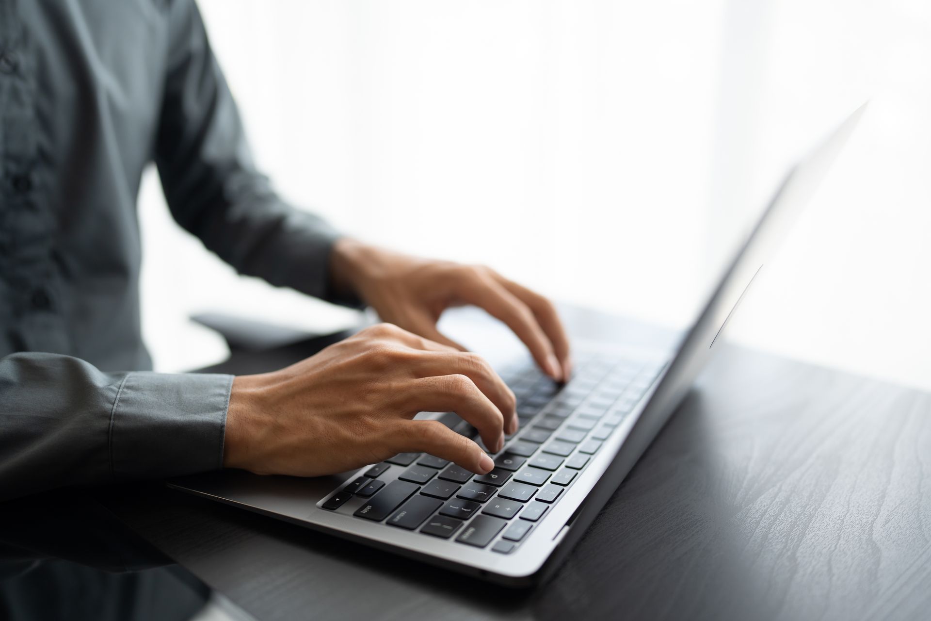 Person typing on a laptop at a desk beside glasses and a small white monitor in a bright workspace