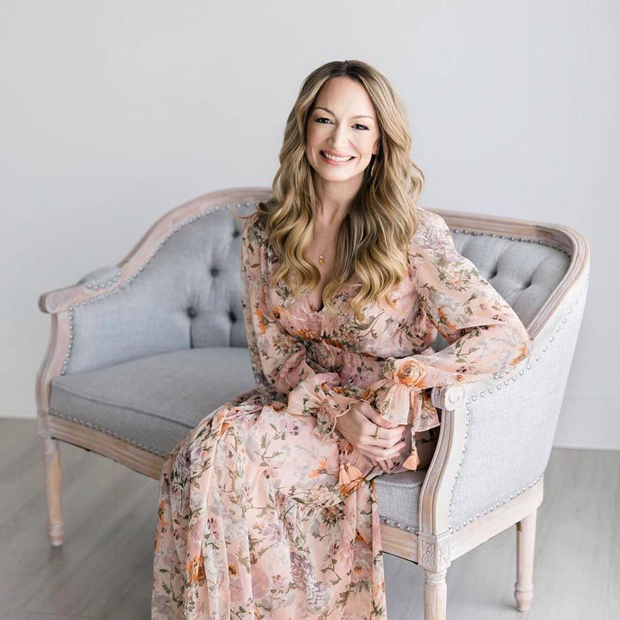 A smiling person with long wavy hair, wearing a floral dress, sits gracefully on a pale blue tufted settee.