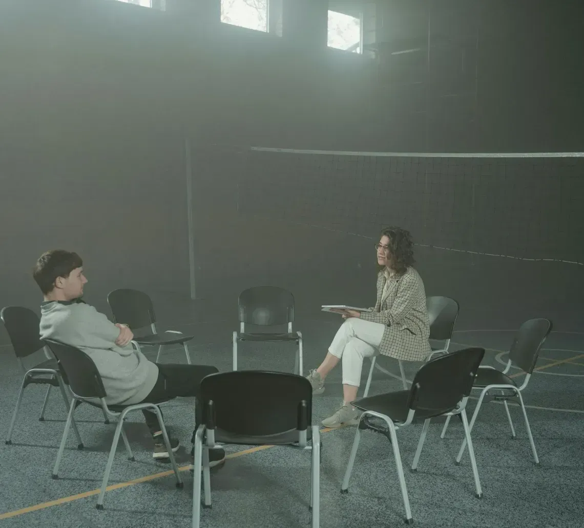 Two people sit in chairs arranged in a circle in a dark, hazy room with high windows, appearing to have a conversation.