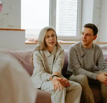A professional session in a bright room where a couple talks to an unseen person while sitting on a soft couch.