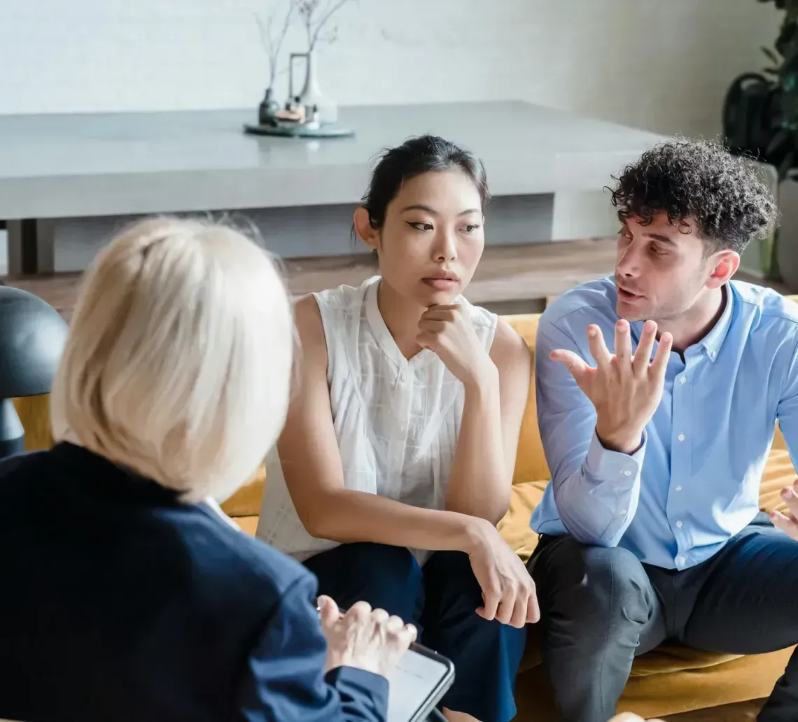 A person in a dark blazer consults with two people seated on a couch; one person gestures while the other listens closely.