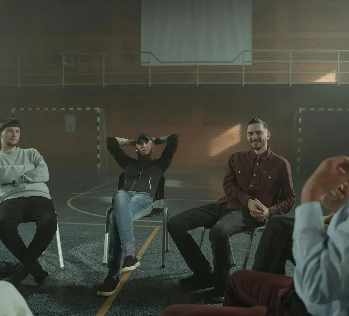 A group of people sit in a circle on chairs in a dimly lit indoor sports court, appearing to be in a discussion.