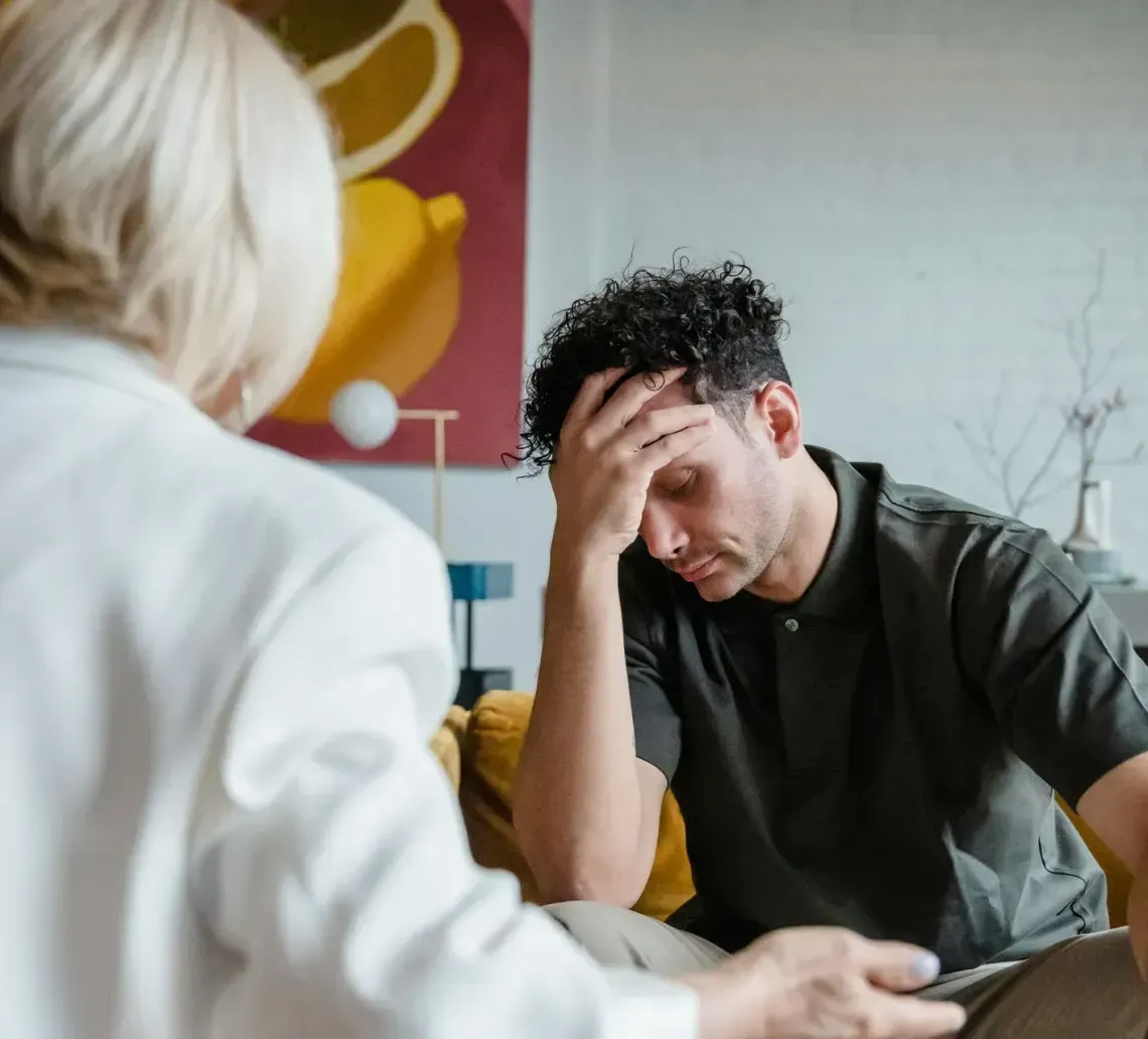 A person sits on a yellow sofa with their hand over their face, facing another person who is listening during a session.
