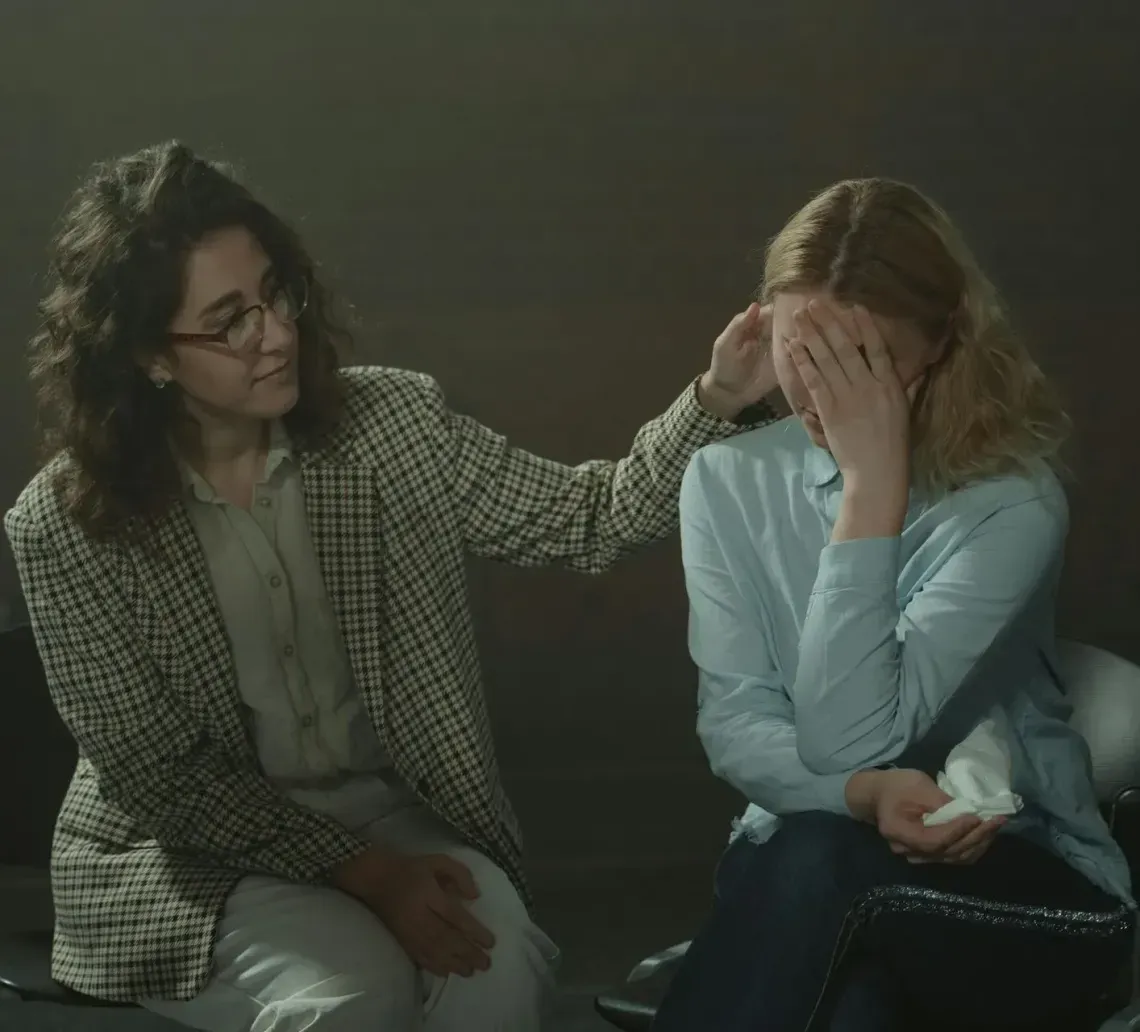 A woman in a plaid blazer comforts another woman, who is covering her face with one hand and holding a tissue.