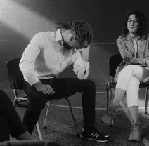 Two people sit on chairs in a gym; one leans forward with their hand on their head, while the other looks on calmly.