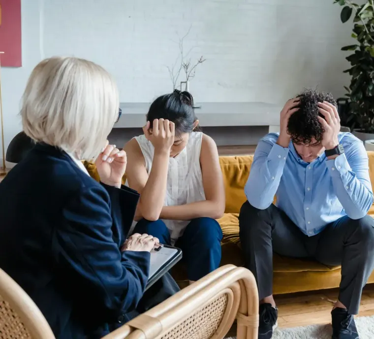 A therapist sits in a chair facing two distressed people sitting on a couch, both with their hands on their heads.