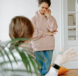 A person in a pink hoodie cries while standing in a room, partially obscured by a foreground plant and a seated person.