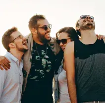 Four friends laugh together, arm-in-arm, outdoors in bright, warm lighting.