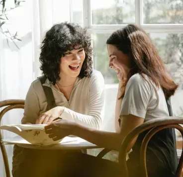 Two people sit at a table by a bright window, laughing and looking down at a book they are reading together.