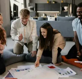 Five people sit on the floor in a bright home, gathered around papers and discussing a project with engaged expressions.