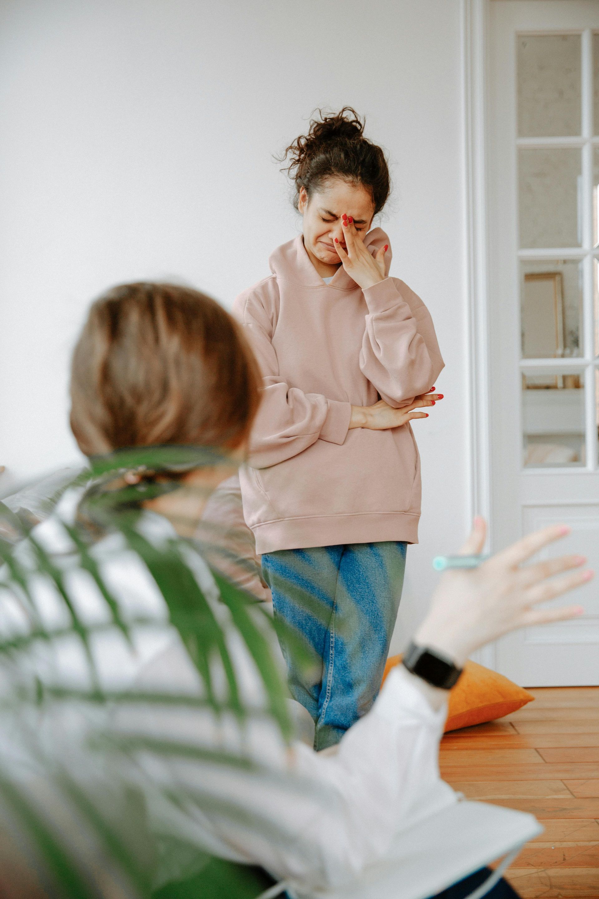 A person in a pink hoodie cries while standing in a room, partially obscured by a foreground plant and a seated person.