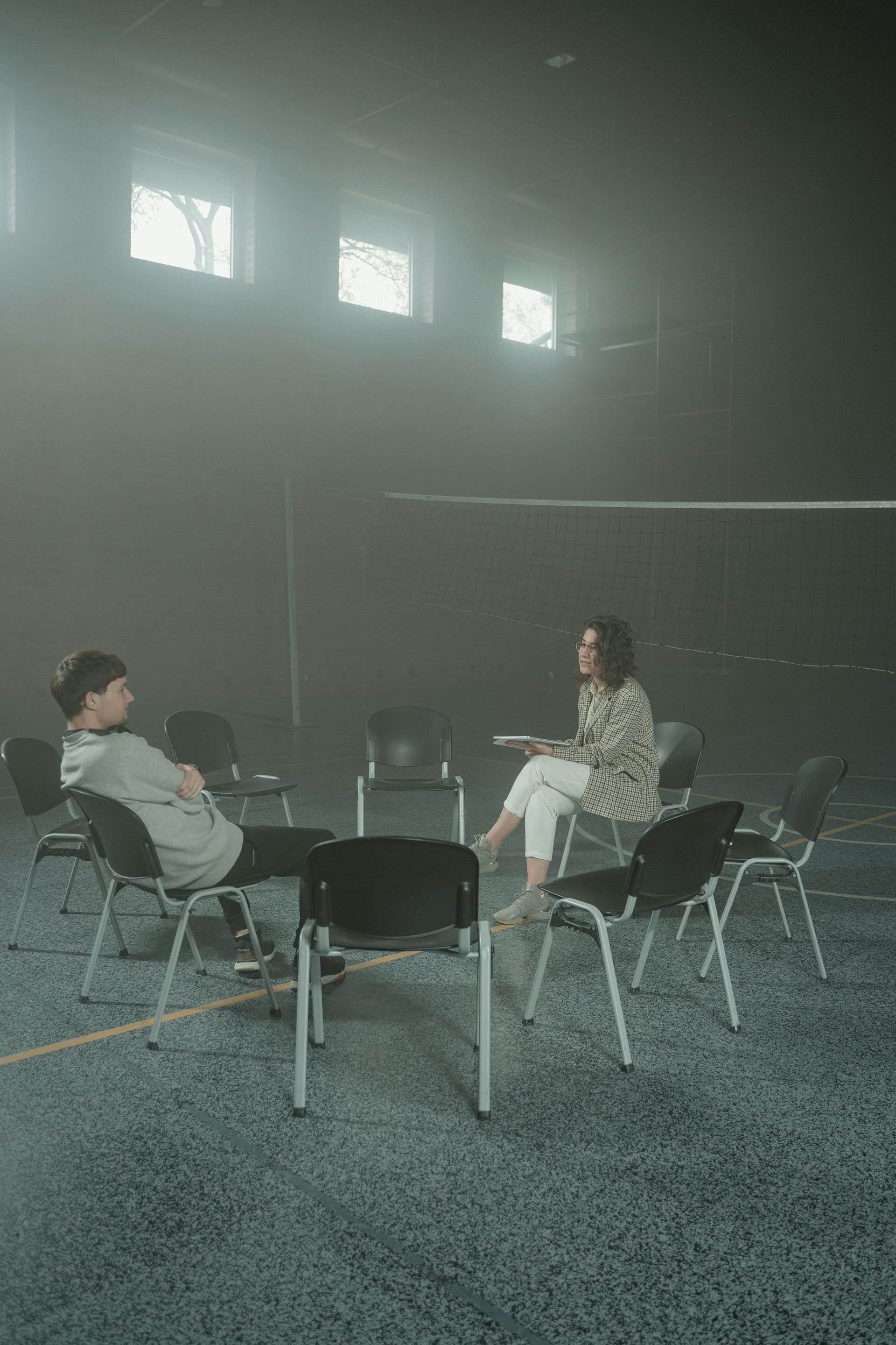Two people sit in chairs arranged in a circle in a dark, hazy room with high windows, appearing to have a conversation.