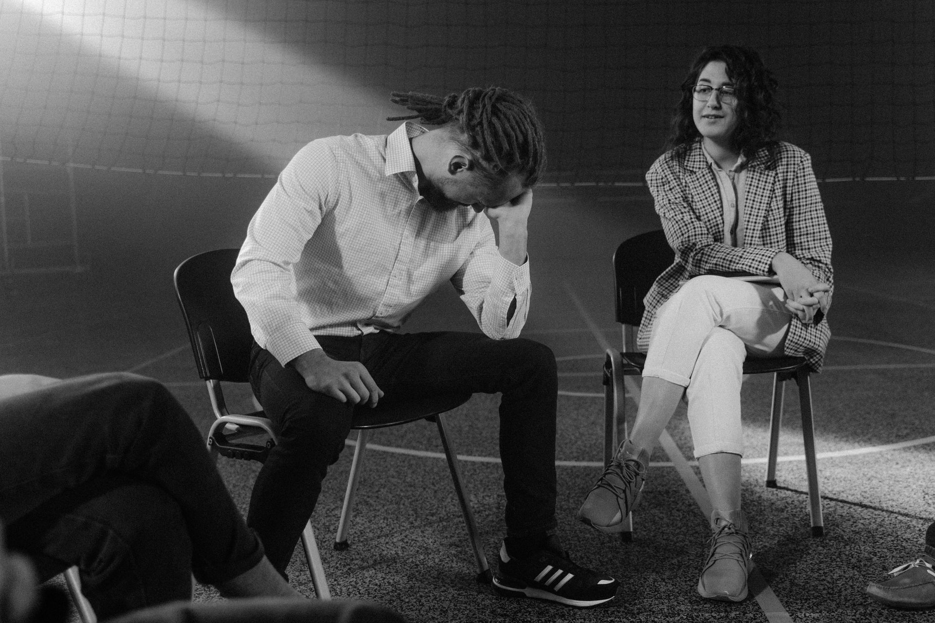 Two people sit on chairs in a gym; one leans forward with their hand on their head, while the other looks on calmly.
