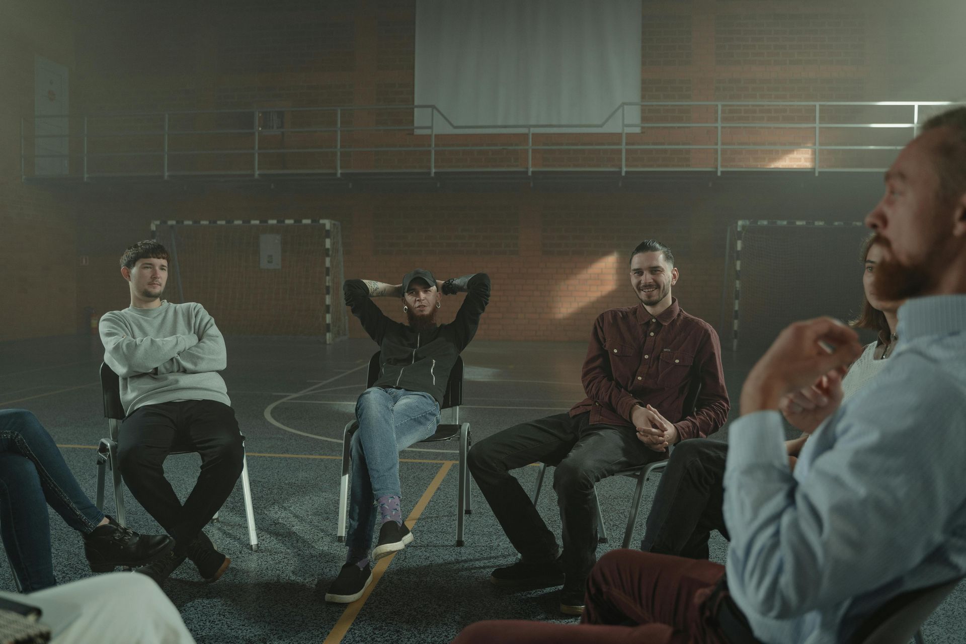 A group of people sit in a circle on chairs in a dimly lit indoor sports court, appearing to be in a discussion.