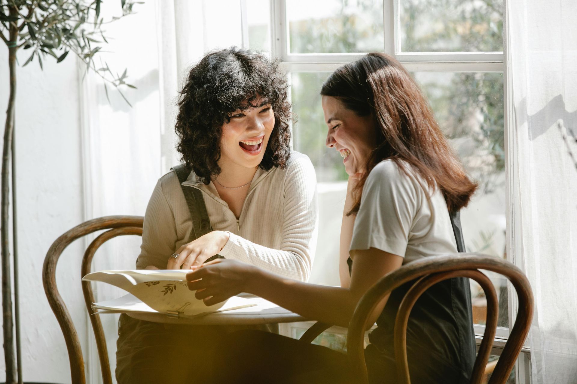 Two people sit at a table by a bright window, laughing and looking down at a book they are reading together.