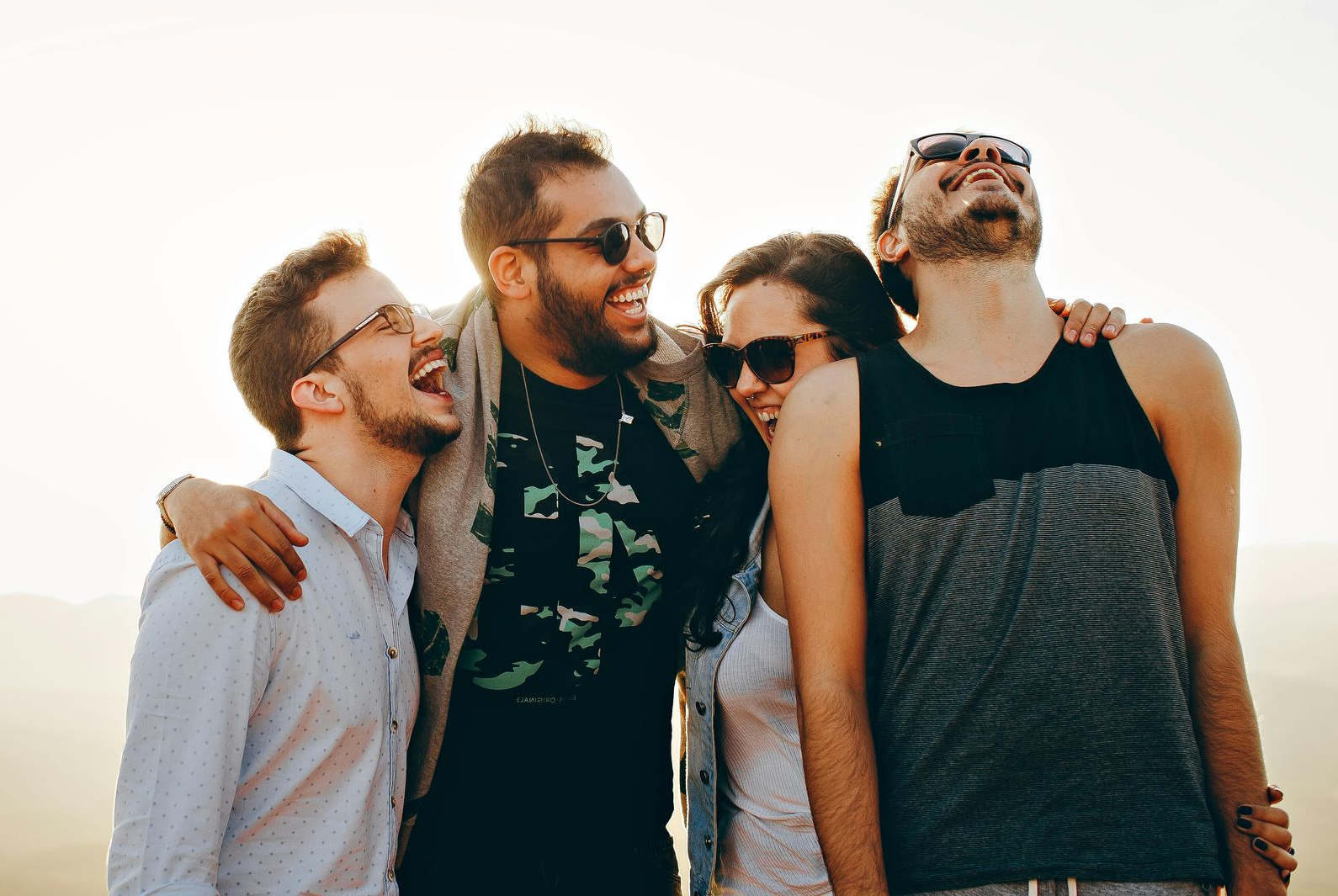 Four friends laugh together, arm-in-arm, outdoors in bright, warm lighting.