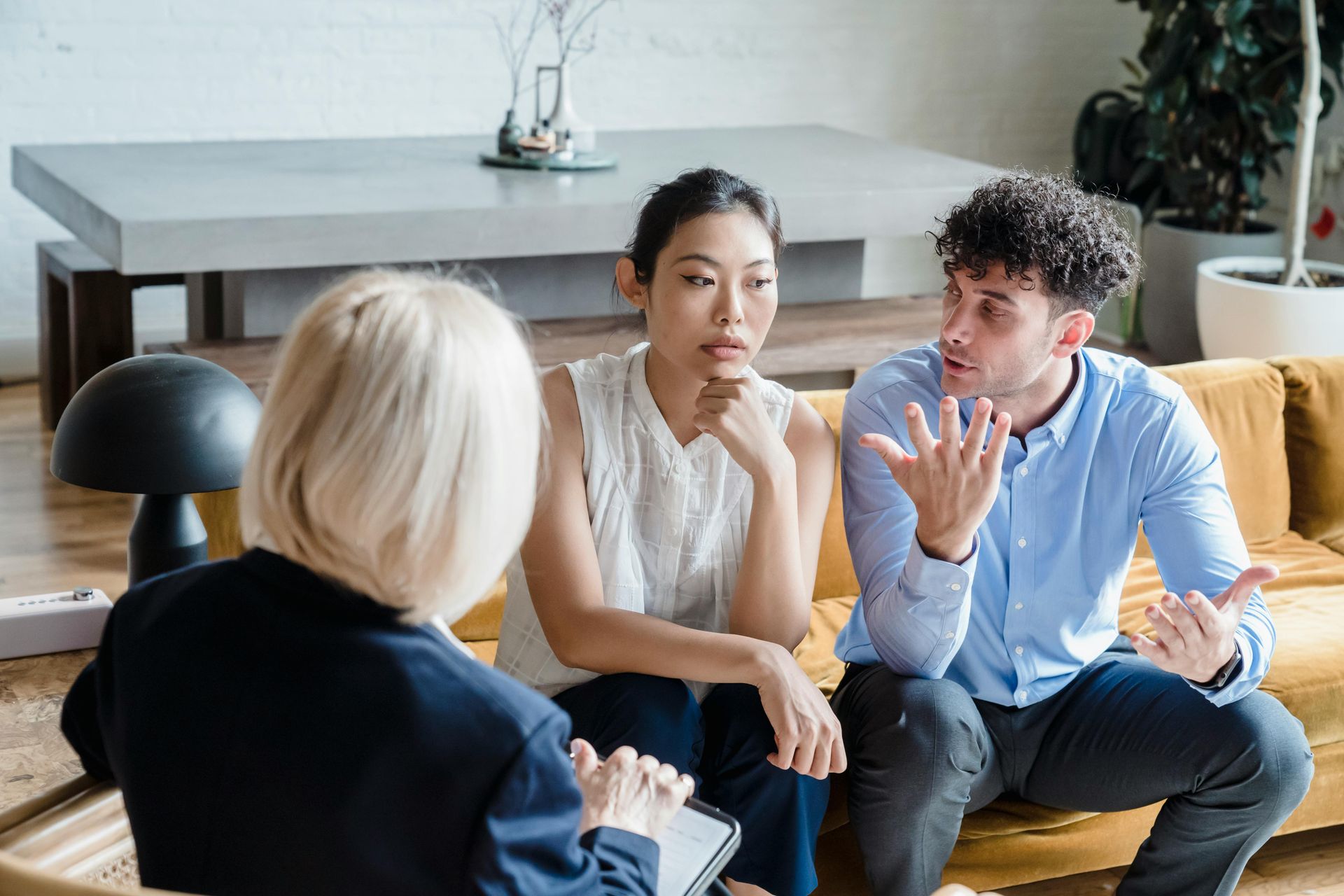 A person in a dark blazer consults with two people seated on a couch; one person gestures while the other listens closely.