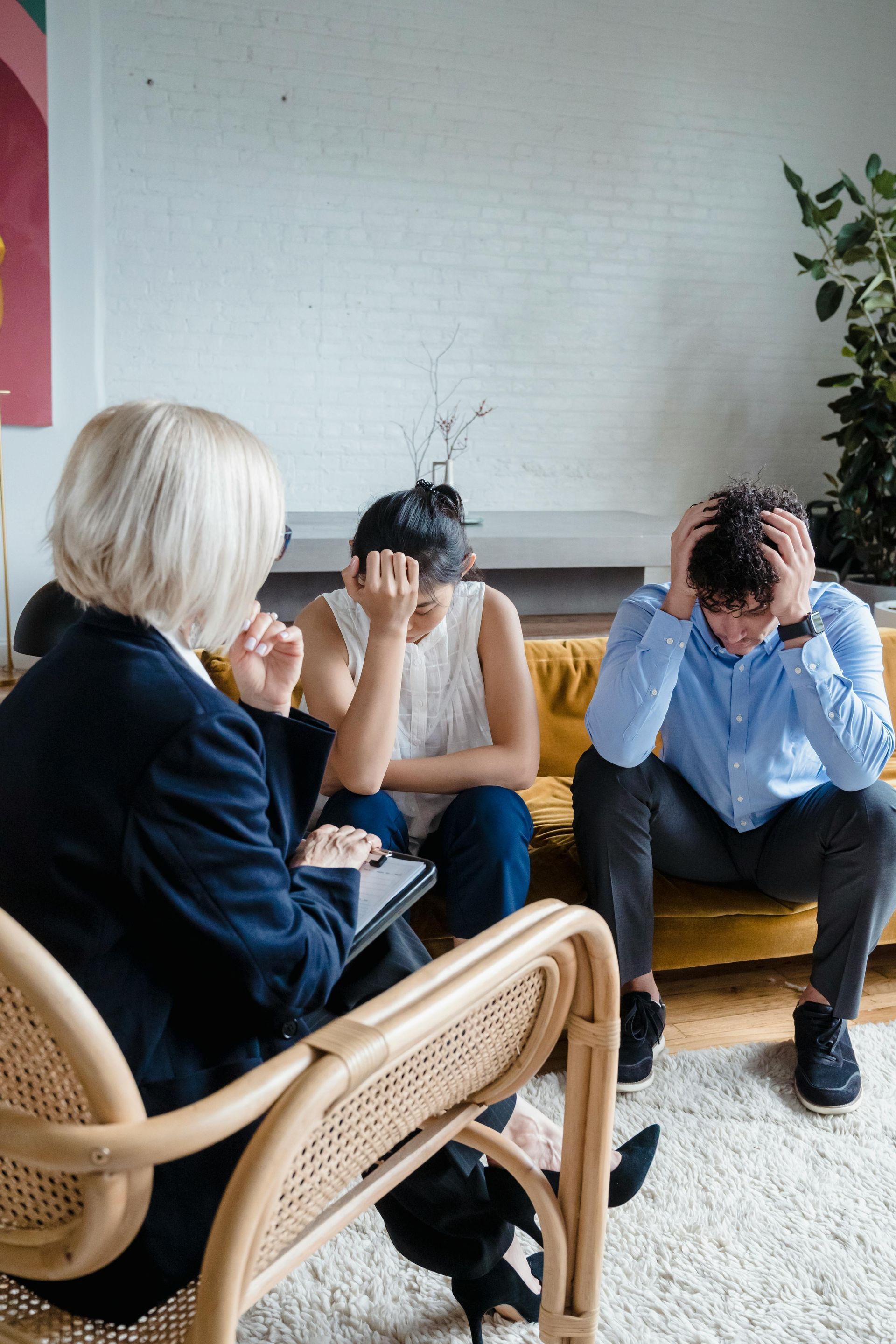 A therapist sits in a chair facing two distressed people sitting on a couch, both with their hands on their heads.