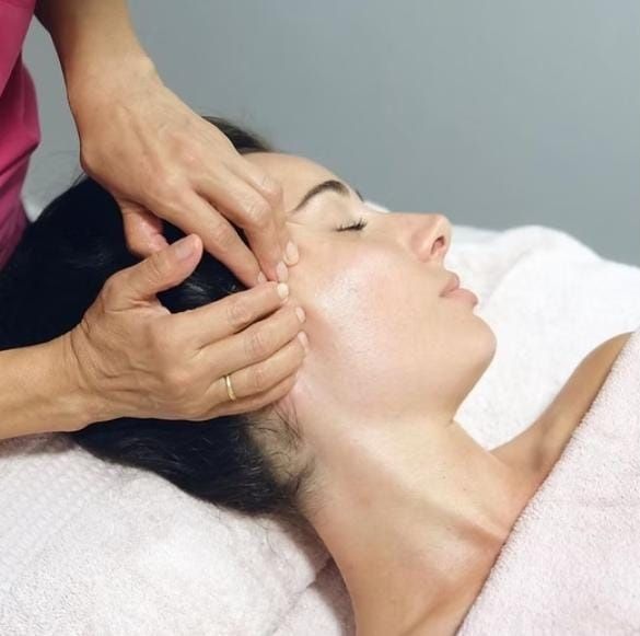 Masaje facial con las manos presionando las sienes y la mejilla de una mujer tumbada en una camilla de tratamiento.