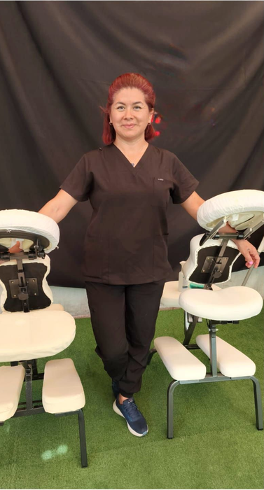 Mujer de pie entre dos sillones de masaje sobre césped artificial, sonriendo a la cámara.