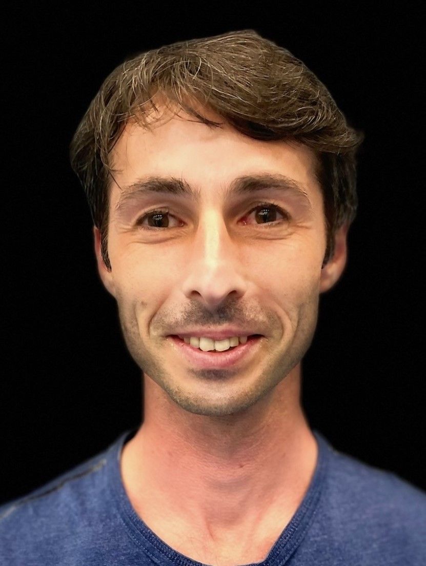 Ein Mann in einem blauen Hemd lächelt vor einem schwarzen Hintergrund.