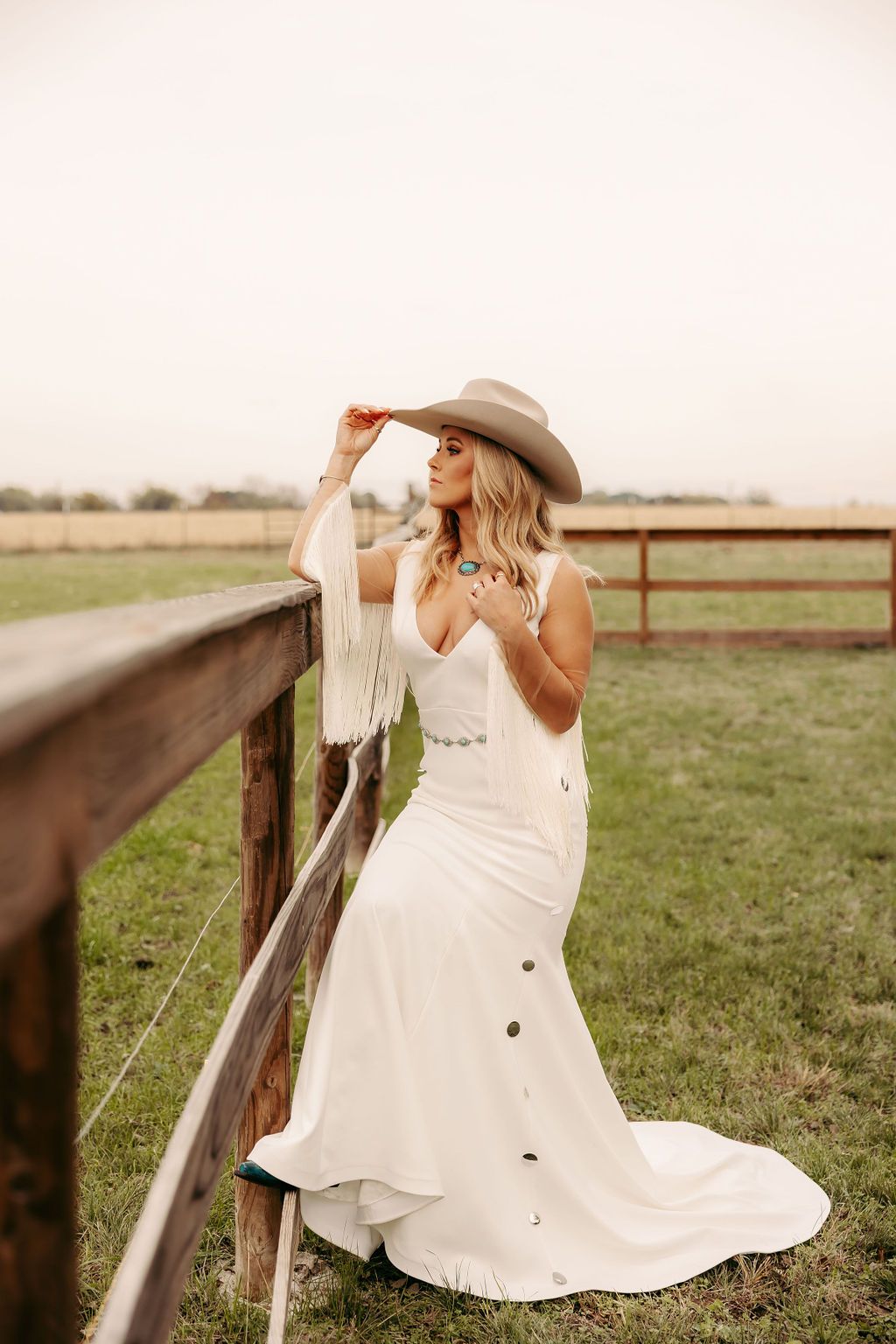 A bride in a white dress and cowboy hat is leaning on a wooden fence.