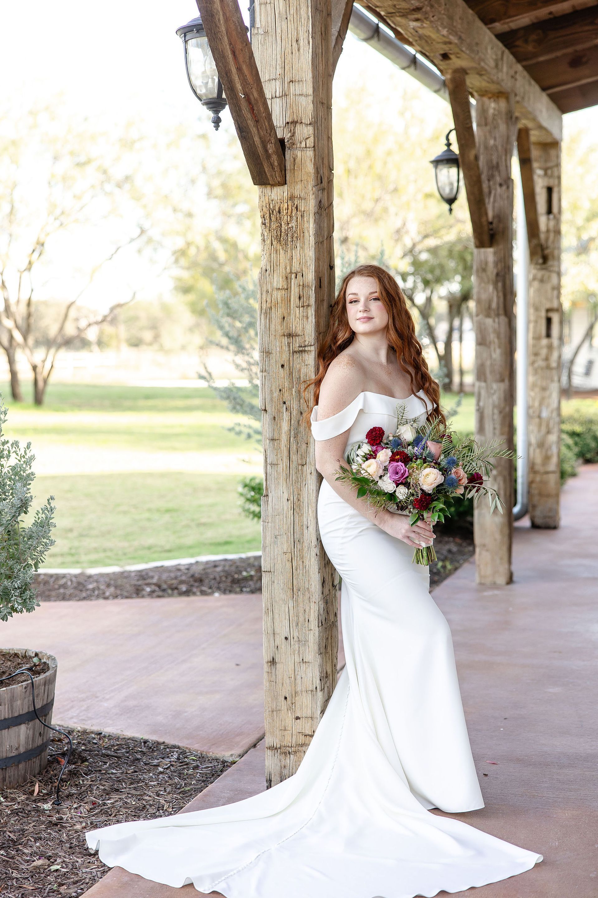 A bride in a white dress is leaning against a wooden post holding a bouquet of flowers.