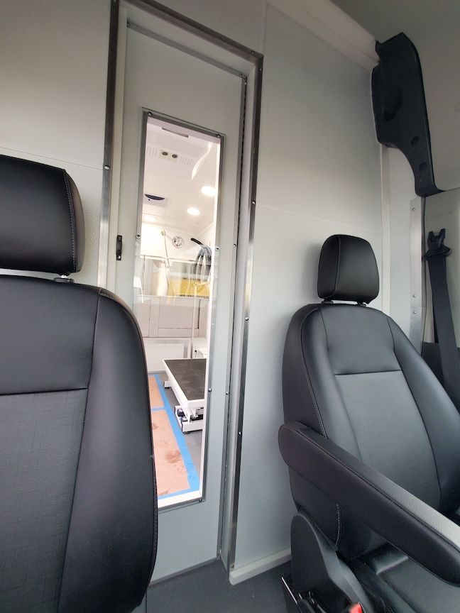 Interior of a white van with black seats and a door leading to a separate room with a grooming table.