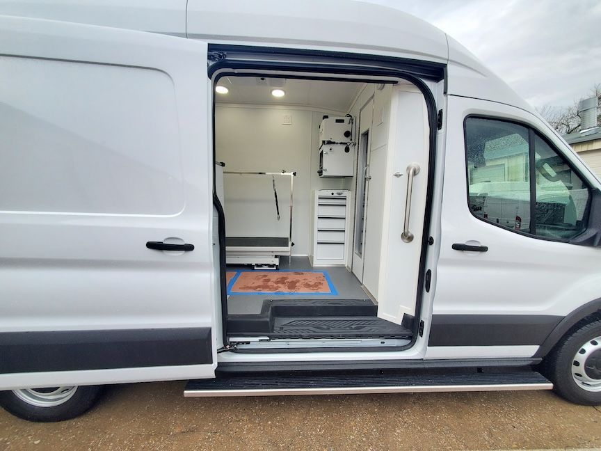 White van with open side door, revealing dog grooming interior, including a grooming table and cabinets.