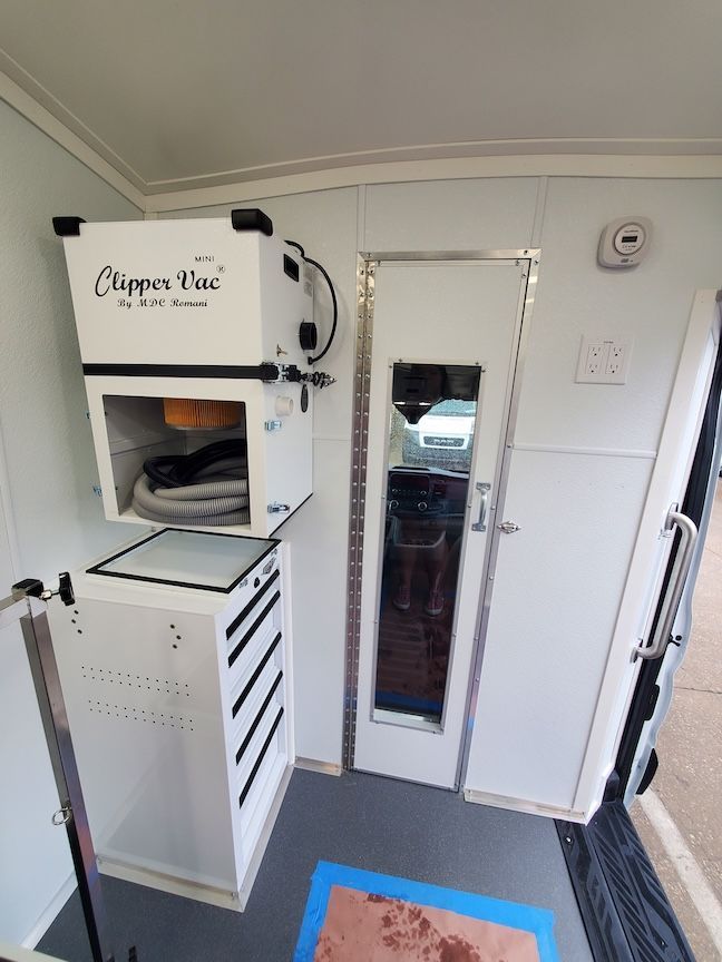 Interior of a white trailer, with a clipper vac system and tool cabinet. A door with a window and a power outlet are also visible.