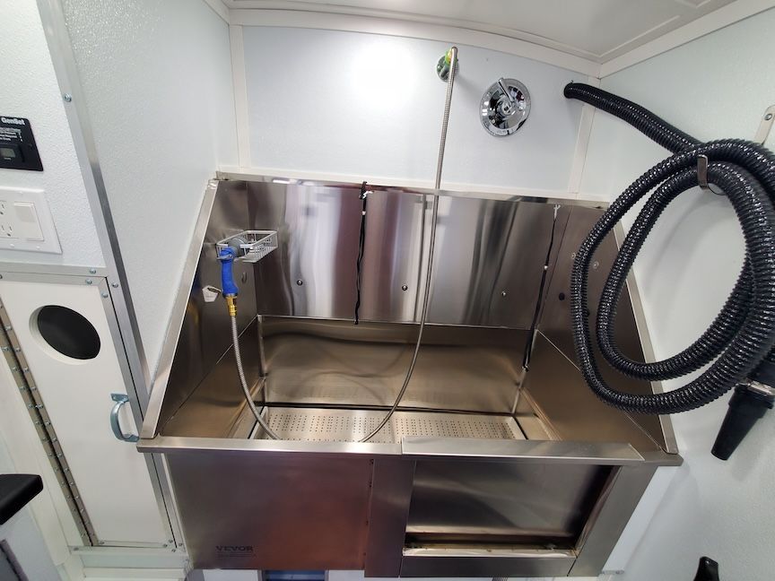 Stainless steel pet grooming tub inside a white-walled trailer, with sprayer and hose.