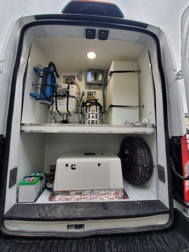 Open back of a white van with medical or industrial equipment. Includes tanks, shelving, a spare tire, and battery.