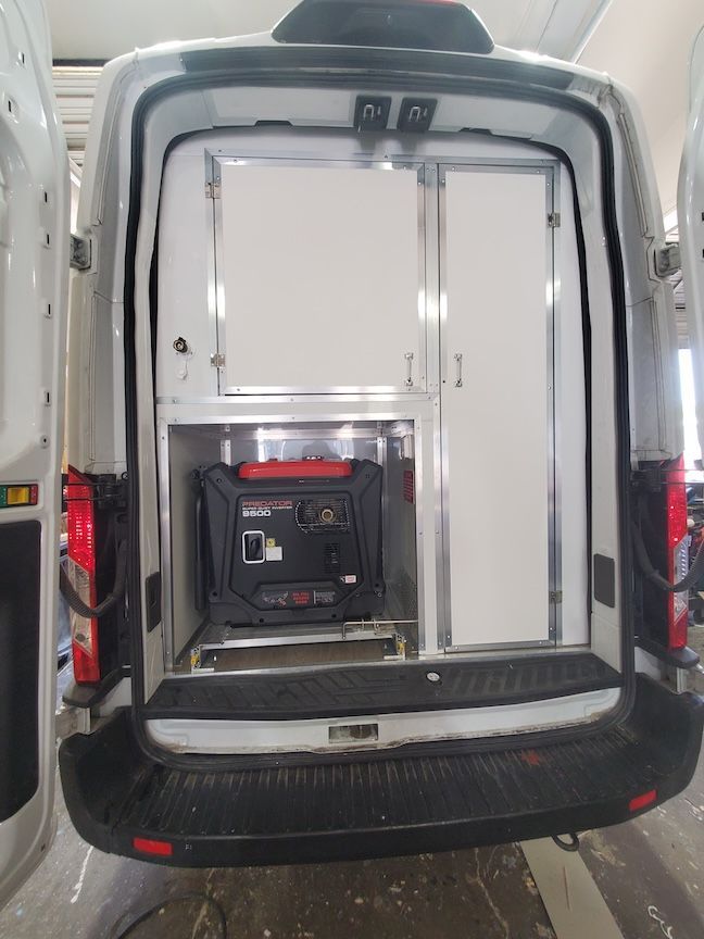 Back of a white van with open doors, showing a generator in a compartment and enclosed cabinet.