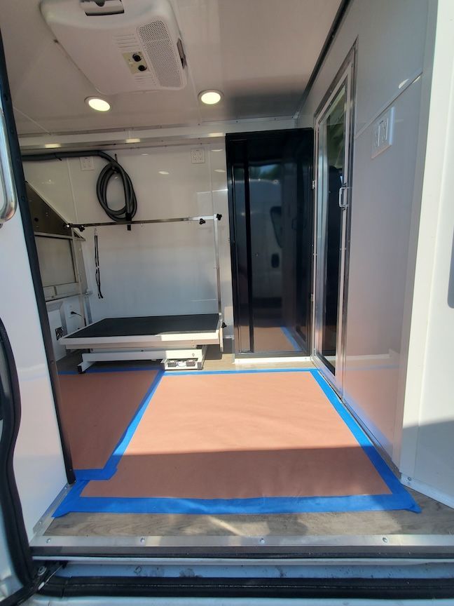 Interior of a white mobile grooming van. Features a grooming table, brown floor, and a doorway.