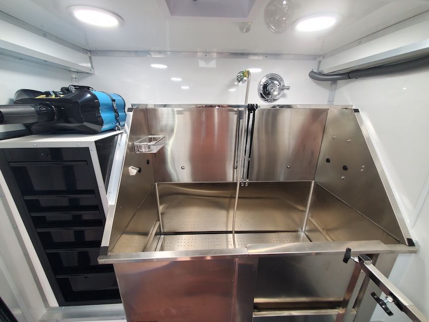 Stainless steel dog wash station inside a grooming trailer, with storage and overhead lighting.