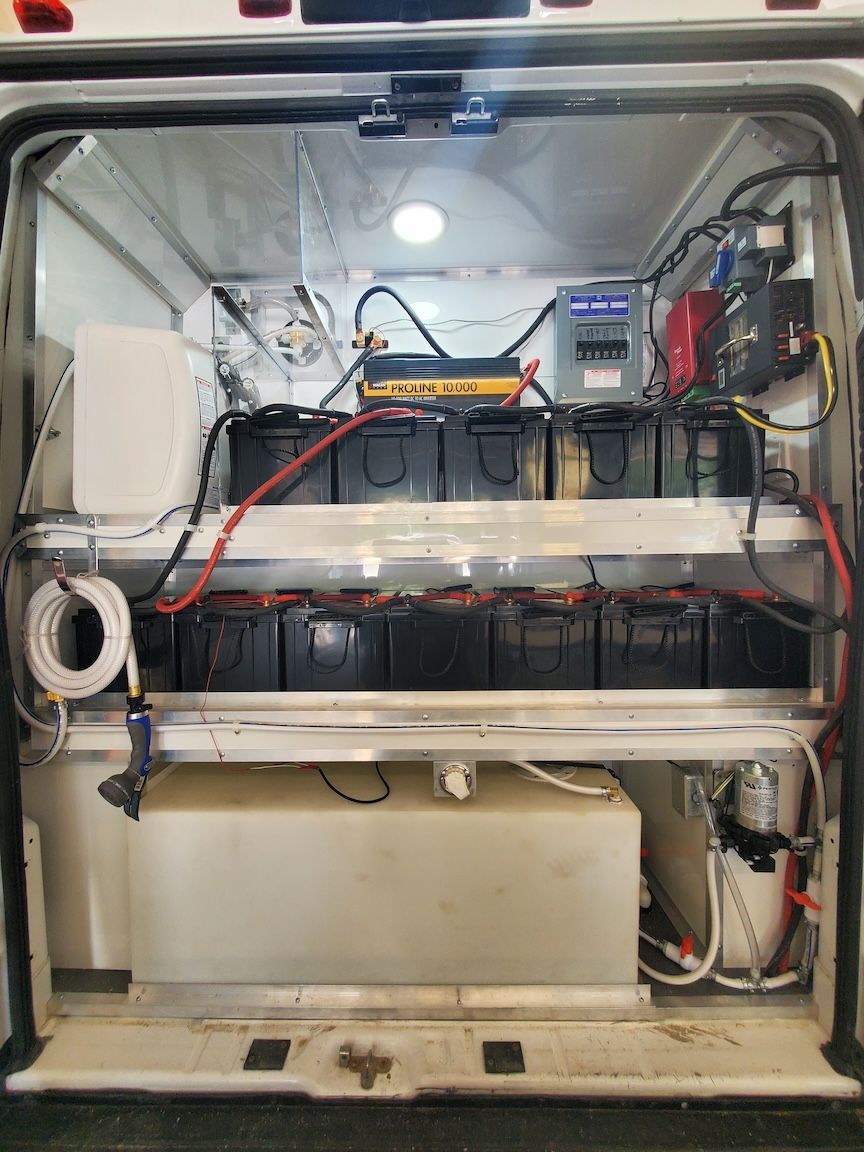 Interior of a vehicle, showing two shelves of batteries, electrical components, and a water tank.