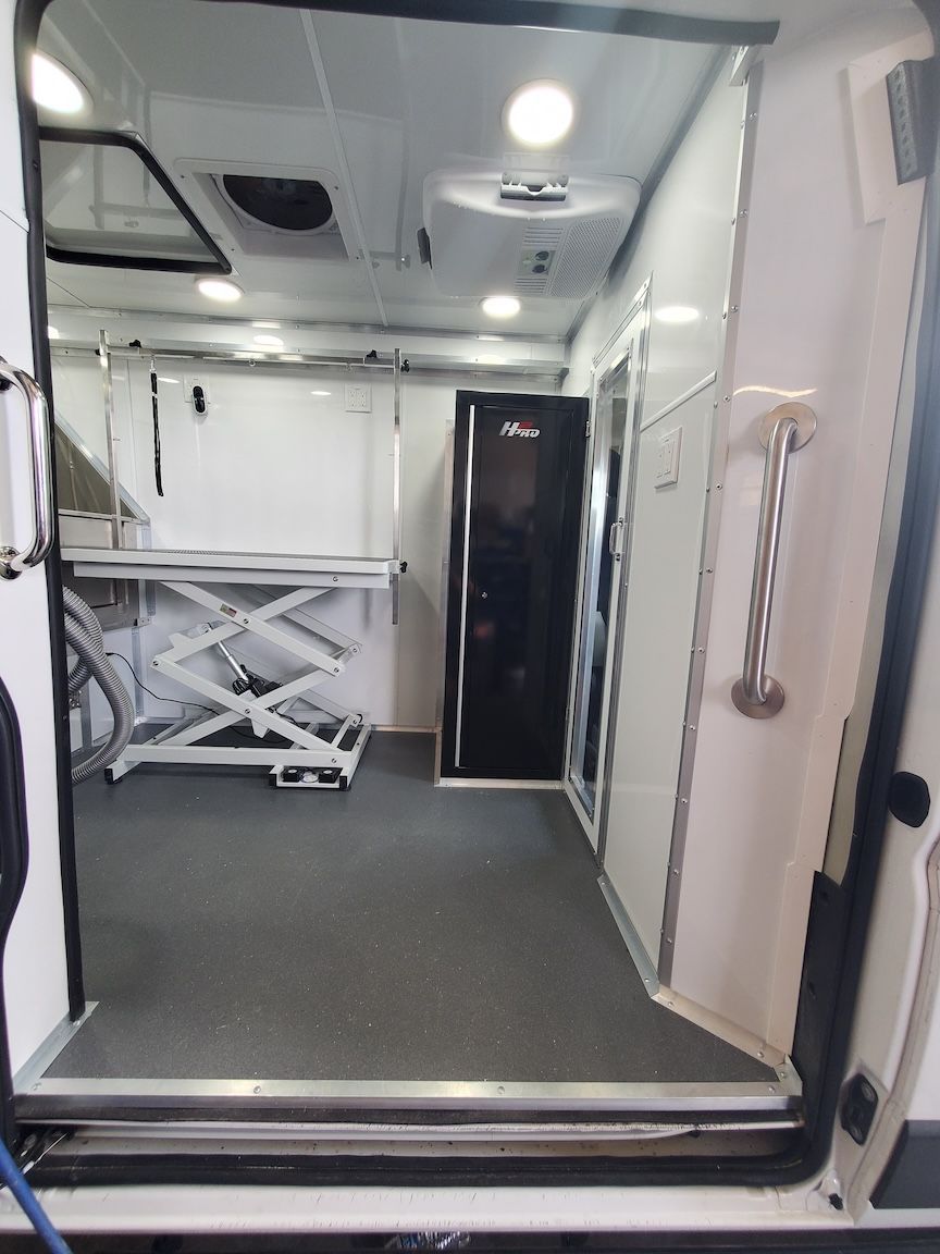 Interior view of a mobile pet grooming van, showing grooming table, storage cabinet, and door. White walls.
