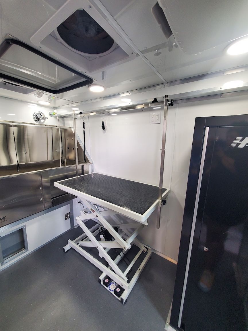 Interior of a pet grooming trailer; a grooming table is in the center, stainless steel cabinets and a vent are visible.