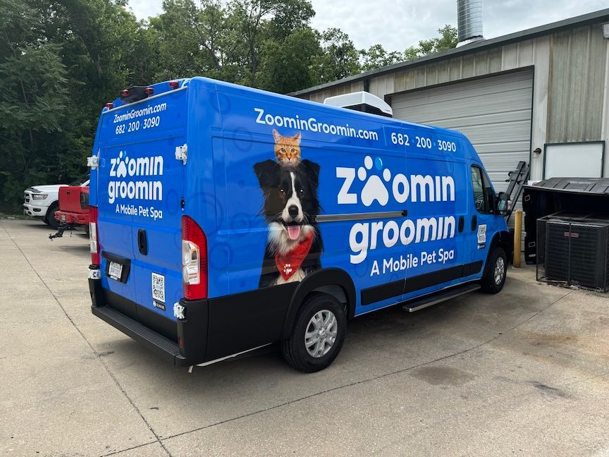 Blue Zoomin' Groomin' mobile pet spa van with dog and cat graphics parked outside a building.