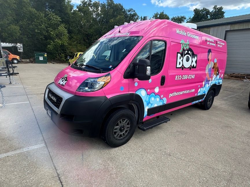 Pink pet grooming van parked outside a building, with logos, phone number, and website.