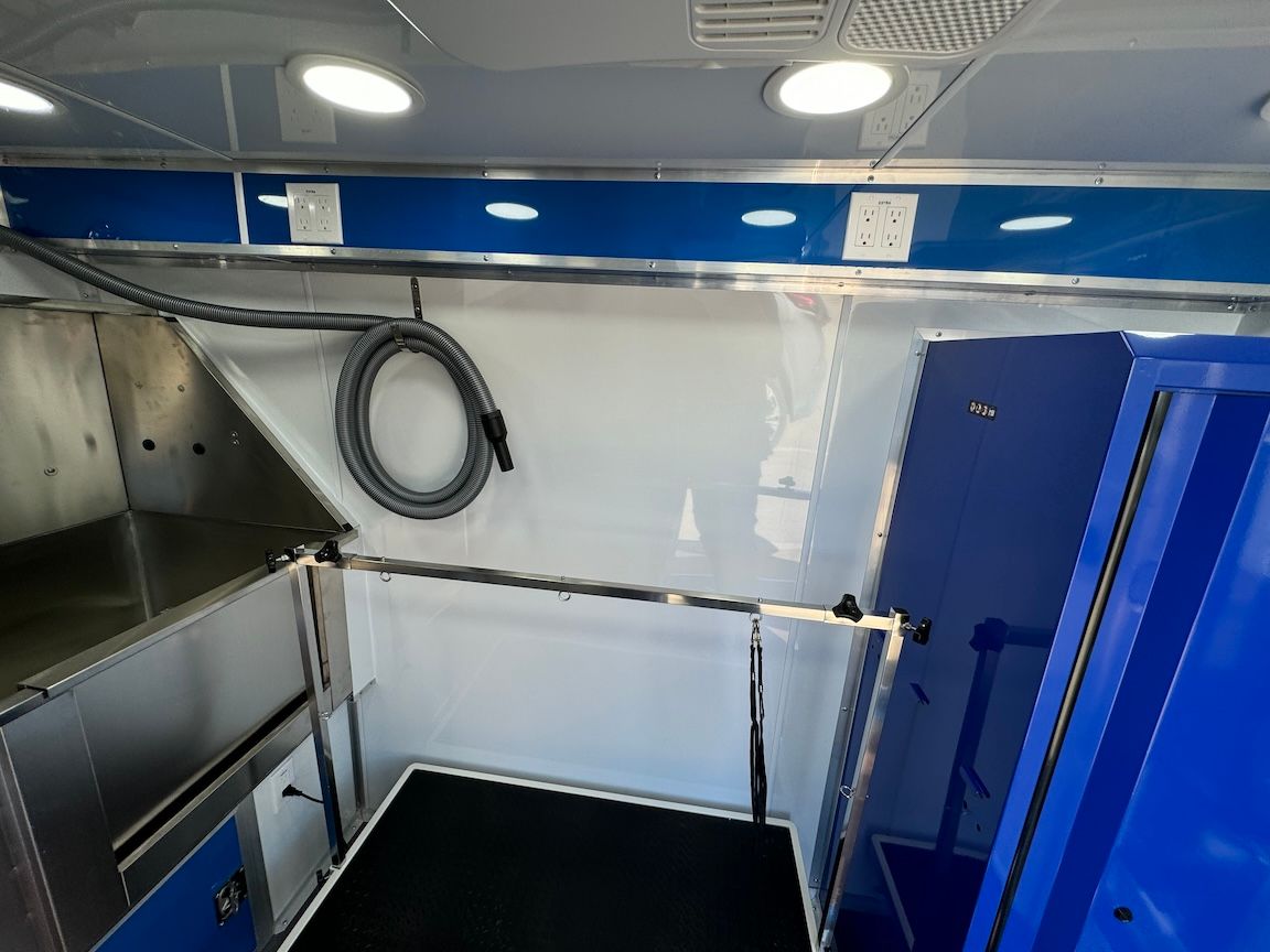 Interior of a mobile pet grooming van: white walls, blue trim, grooming table, vacuum hose, and lighting.