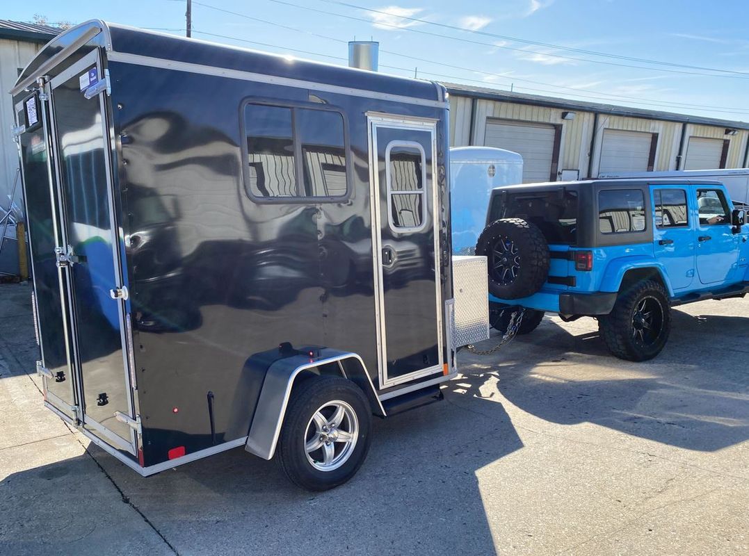 Black trailer hitched to a blue Jeep. Trailer has windows, Jeep has spare tire.