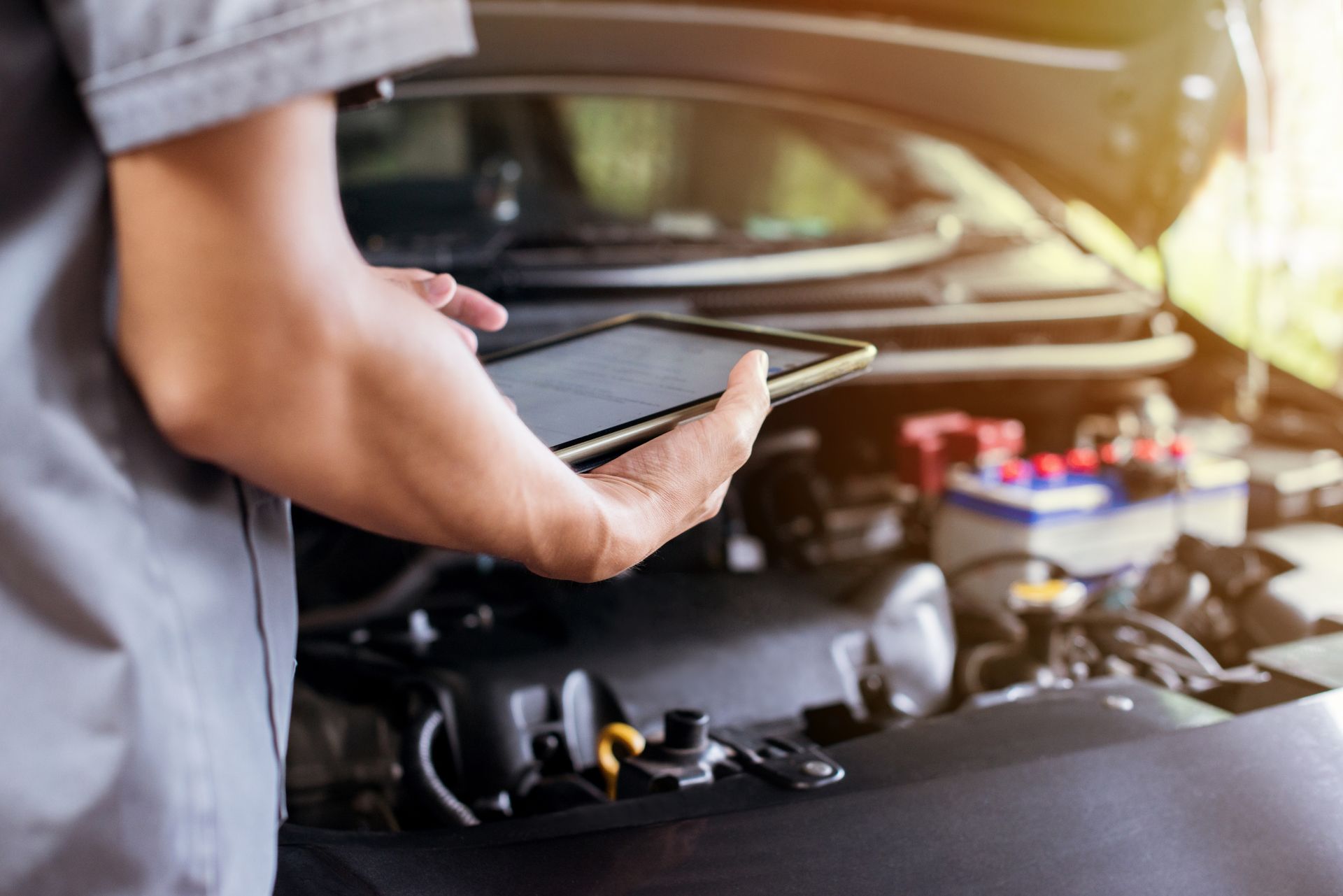 A mechanic in a grey uniform holds a tablet while inspecting an open car engine.