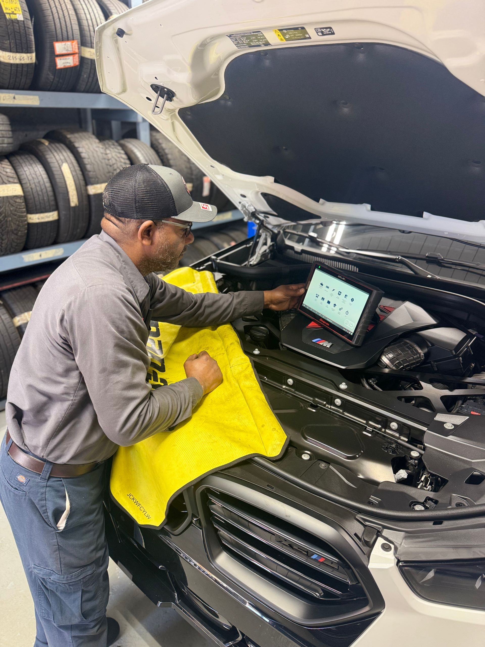 Mechanic using a diagnostic tool on a car in a shop. He is wearing a grey shirt, hat and jeans.