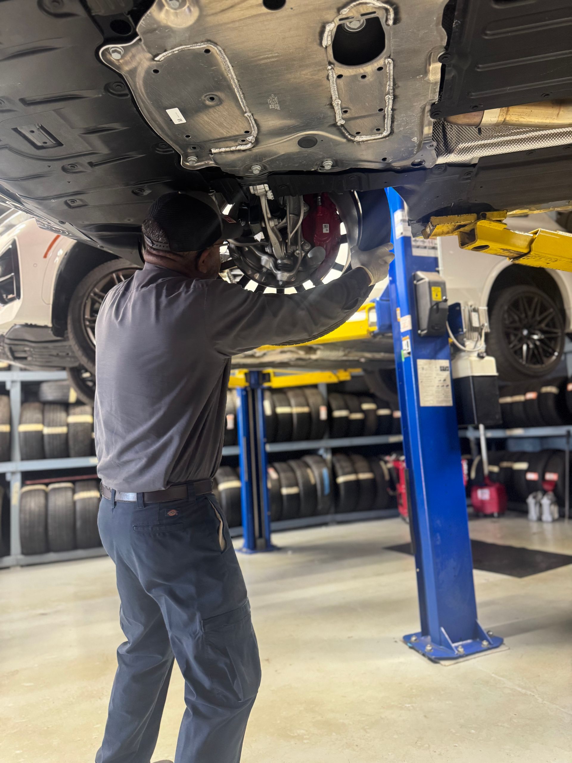 Mechanic working under a car on a lift in a shop, wearing gray shirt and jeans. Tires are in the background.