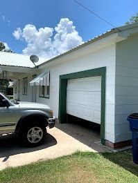 A car is parked in front of a white house with a green garage door.